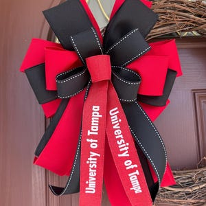 May include: A decorative bow featuring red and black ribbons with white stitching. Two red ribbons with the text "University of Tampa" are centered. The bow is likely for a wreath or door decoration.