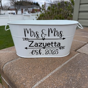 May include: White metal oval-shaped container with black lettering that reads "Mrs. & Mrs. Zazuetta est. 2025" with arrows and a heart.