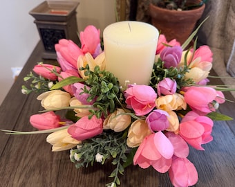 Peut inclure: Une bougie blanche en forme de pilier entourée d'un arrangement floral de tulipes roses, violettes et crème avec un feuillage vert.