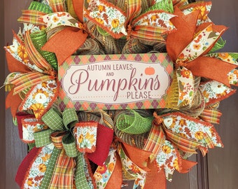 May include: A fall-themed wreath with orange, green, and brown ribbons, burlap, and mesh. The wreath has a sign that says "Autumn Leaves and Pumpkins Please".