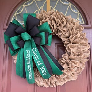 May include: A burlap wreath with a large bow featuring green and black ribbons. The bow has the text "Norman North High School" and "Class of 2026" in white. The wreath is hanging on a brown door.