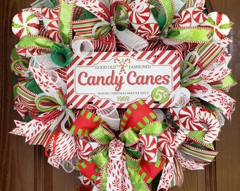 May include: A red, white, and green Christmas wreath with a sign that reads "Good Old Fashioned Candy Canes Making Christmas Sweeter Since 1902 5c". The wreath is decorated with candy cane patterned ribbon, green and red bows, and white mesh.
