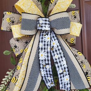May include: A yellow and black bee-themed door bow with a burlap base and a black and white gingham ribbon. The bow is decorated with yellow and white floral print ribbon and black polka dot ribbon.