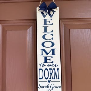 May include: A vertical rectangular white wooden sign with dark blue text reading WELCOME to our DORM, followed by the names Sarah Grace & Elle. The sign features a dark blue fabric bow at the top and is suspended by a piece of brown twine.