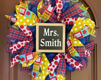 May include: A decorative wreath featuring a central square sign with the text Mrs. Smith. The wreath is constructed from blue mesh and accented with ribbons in yellow with white polka dots, red with white polka dots, blue plaid, and school-themed prints.