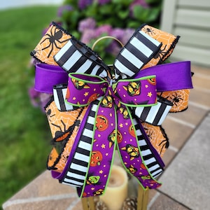 May include: A Halloween-themed bow with multiple layers of ribbon. The bow features orange spiderweb and black spider print ribbon, black and white striped ribbon, and purple ribbon. A central ribbon displays jack-o'-lanterns and black cats. The bow is placed on a small lantern.