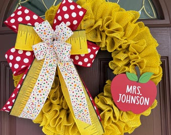 May include: A handmade yellow burlap wreath with a red and white polka dot bow and a bow with a ruler design. A red apple-shaped sign with the text "MRS. JOHNSON" is attached to the wreath. The wreath is hanging on a brown door.
