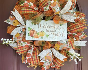May include: A fall-themed wreath with a variety of ribbons in orange, green, and plaid patterns. The wreath features a sign that reads "Welcome to our patch" with pumpkin illustrations. White floral accents are also included.