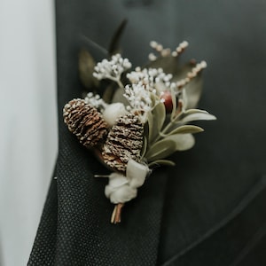 May include: A brown and white floral buttonhole with pine cones and greenery.