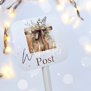 May include: A white sign with a watercolor illustration of a wrapped gift box with a brown bow and greenery. The sign reads "Weihnachts Post" in gold script and block letters. The sign is on a white stick.
