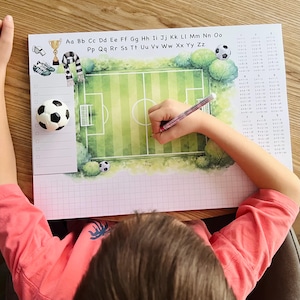 Könnte beinhalten: Eine rechteckige Schreibtischunterlage mit einer Aquarell-Illustration eines Fußballfeldes, dem Alphabet von Aa bis Zz, einem Einmaleins, einer Checkliste und fußballbezogenen Symbolen wie einem Pokal, einer Pfeife und einem Fußball.
