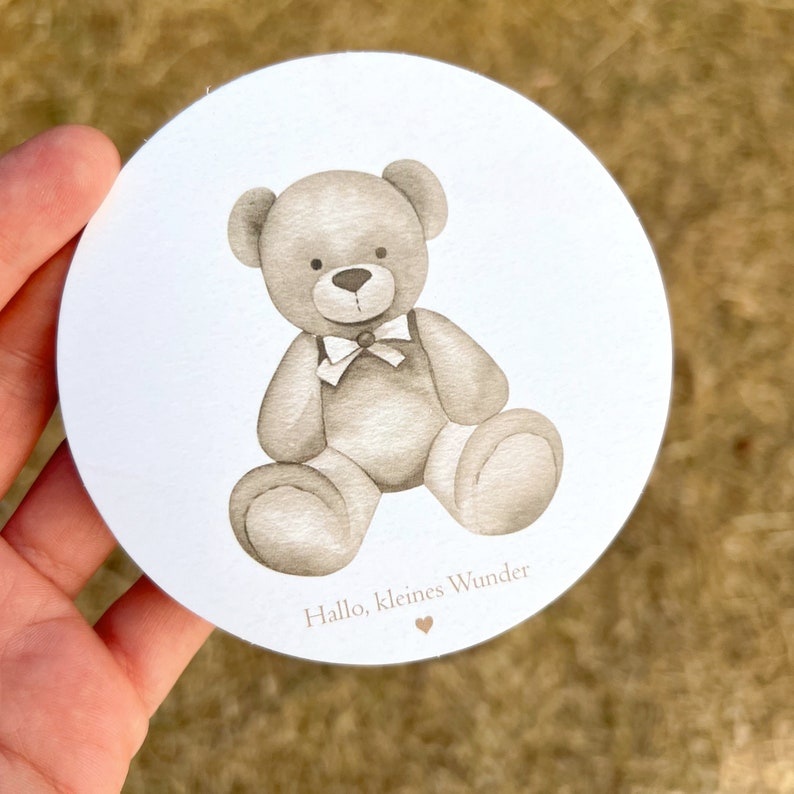 Op de afbeelding: Een ronde witte kaart met een bruine teddybeer met een strikje. De tekst "Hallo, kleines Wunder" staat op de kaart gedrukt.
