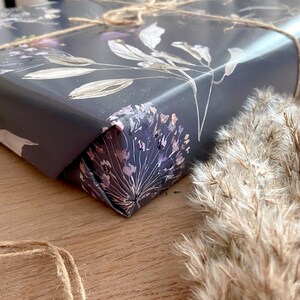 May include: Close-up of a corner of a gift wrapped in dark grey wrapping paper with a floral pattern in shades of grey, white, and purple. The paper is tied with twine.