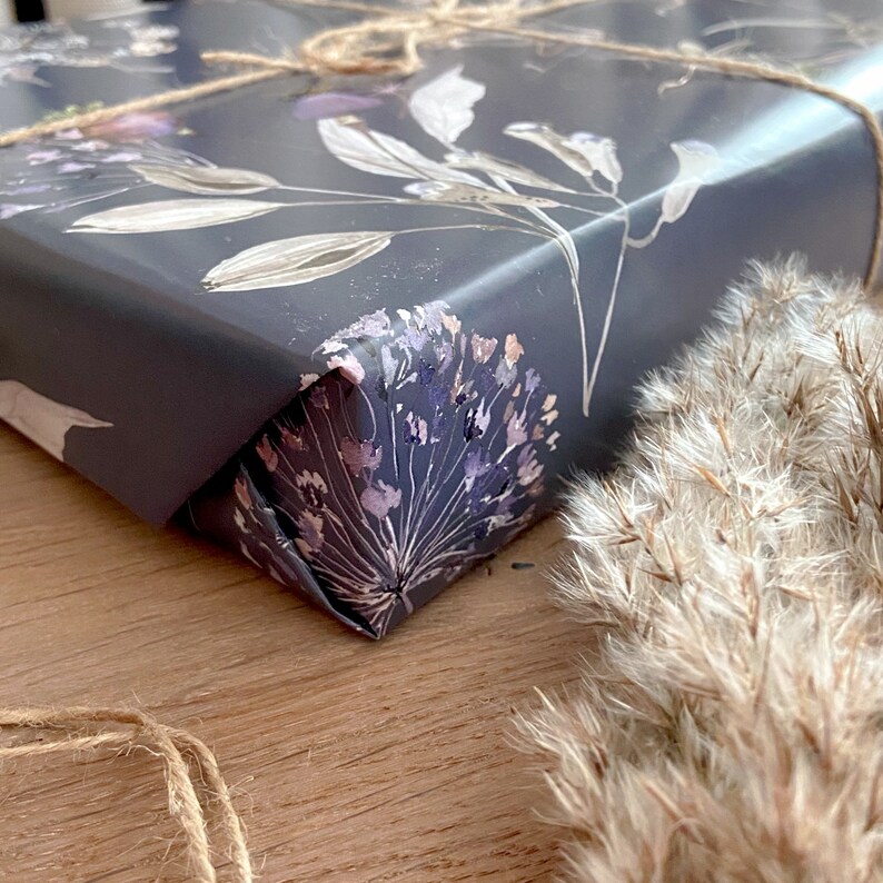 May include: Close-up of a corner of a gift wrapped in dark grey wrapping paper with a floral pattern in shades of grey, white, and purple. The paper is tied with twine.