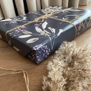 May include: Dark grey gift wrap with a floral pattern featuring dried flowers and leaves. The wrapping paper is tied with twine.
