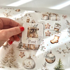 May include: A hand holding a square sticker with the text "Warmont's Post!" over Christmas-themed wrapping paper. The wrapping paper features illustrations of gifts, ornaments, gingerbread stars, and a house with a chimney, all in neutral tones.