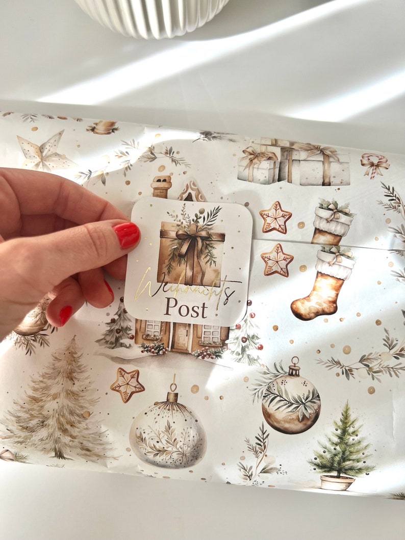 May include: A hand holding a square sticker with the text "Warmont's Post!" over Christmas-themed wrapping paper. The wrapping paper features illustrations of gifts, ornaments, gingerbread stars, and a house with a chimney, all in neutral tones.