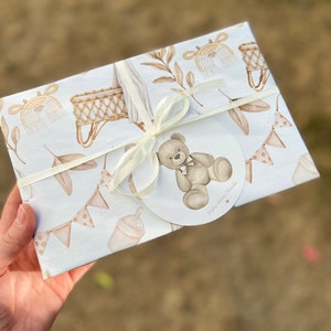 Op de afbeelding: Witte cadeaupapier met een bruine teddybeer, een wieg en een fles. Het cadeaupapier heeft een wit lint en een label met de tekst "Hello Little Wonder".