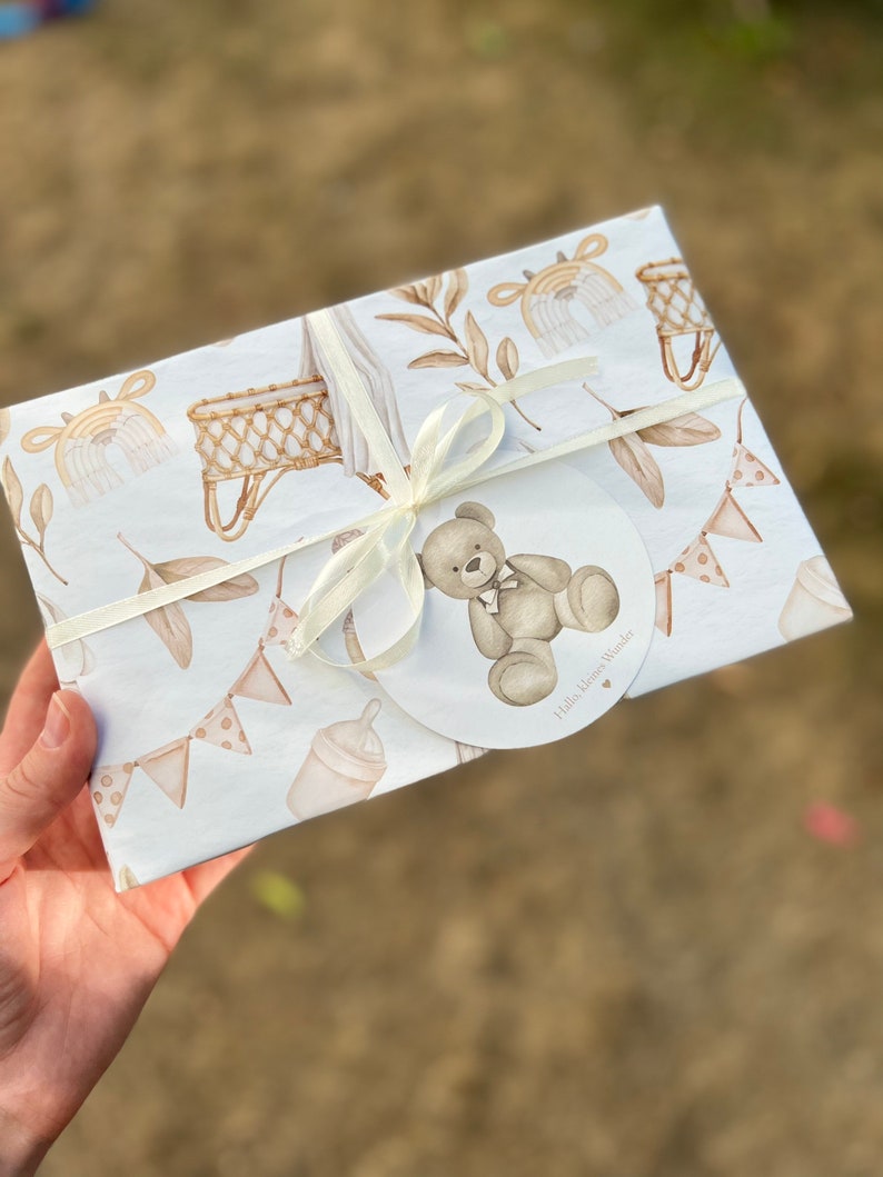 Op de afbeelding: Witte cadeaupapier met een bruine teddybeer, een wieg en een fles. Het cadeaupapier heeft een wit lint en een label met de tekst "Hello Little Wonder".