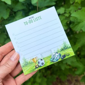 Könnte beinhalten: Ein quadratischer Notizblock mit dem Text MEINE TO-DO LISTE am oberen Rand, der fünf linierte Zeilen mit runden Checkboxen enthält. Der untere Bereich zeigt eine Aquarellillustration eines Fußballfeldes, einen Pokal, einen Ball und eine Pfeife.