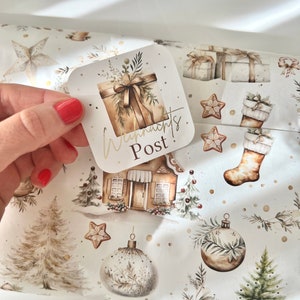 May include: A square gift tag with the words "Weihnachts Post" in gold lettering is held over Christmas-themed wrapping paper. The wrapping paper features illustrations of gifts, ornaments, gingerbread houses, and Christmas trees in a neutral color palette.