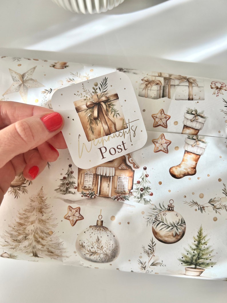 May include: A square gift tag with the words "Weihnachts Post" in gold lettering is held over Christmas-themed wrapping paper. The wrapping paper features illustrations of gifts, ornaments, gingerbread houses, and Christmas trees in a neutral color palette.