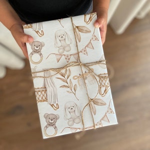 Op de afbeelding: Wit cadeaupapier met een bruin en beige patroon van teddyberen, puppy's en andere babythema-illustraties. Het cadeaupapier is vastgebonden met touw.