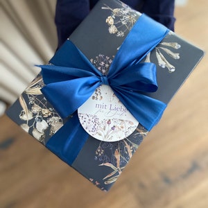 May include: A dark blue gift box with a blue satin ribbon and a white circular tag that says "mit Liebe f&uuml;r dich". The box is wrapped in paper with a floral pattern.