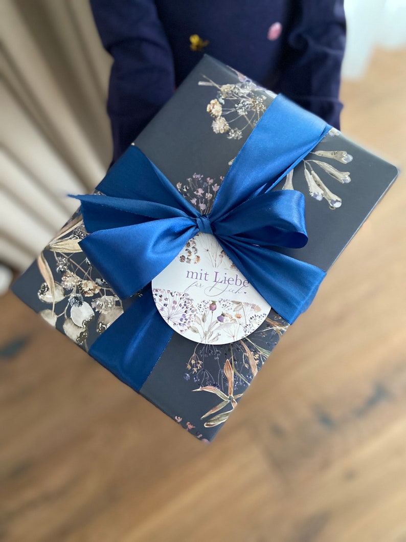 May include: A dark blue gift box with a blue satin ribbon and a white circular tag that says "mit Liebe f&uuml;r dich". The box is wrapped in paper with a floral pattern.