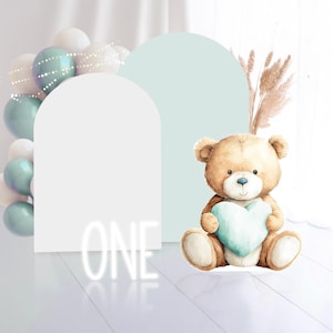 Può includere: Un orsacchiotto marrone chiaro con un cuore azzurro chiaro, posto davanti ad archi bianchi e una ghirlanda di palloncini. La parola "ONE" è illuminata al neon bianco. La scena ha una palette di colori pastello.