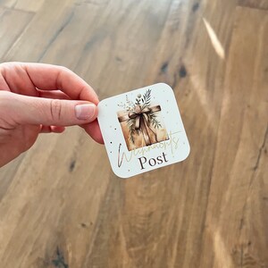 May include: A square sticker with rounded corners featuring a watercolor illustration of a wrapped gift box with a brown bow and greenery. The sticker has the words "Wynn's Post" in gold script.