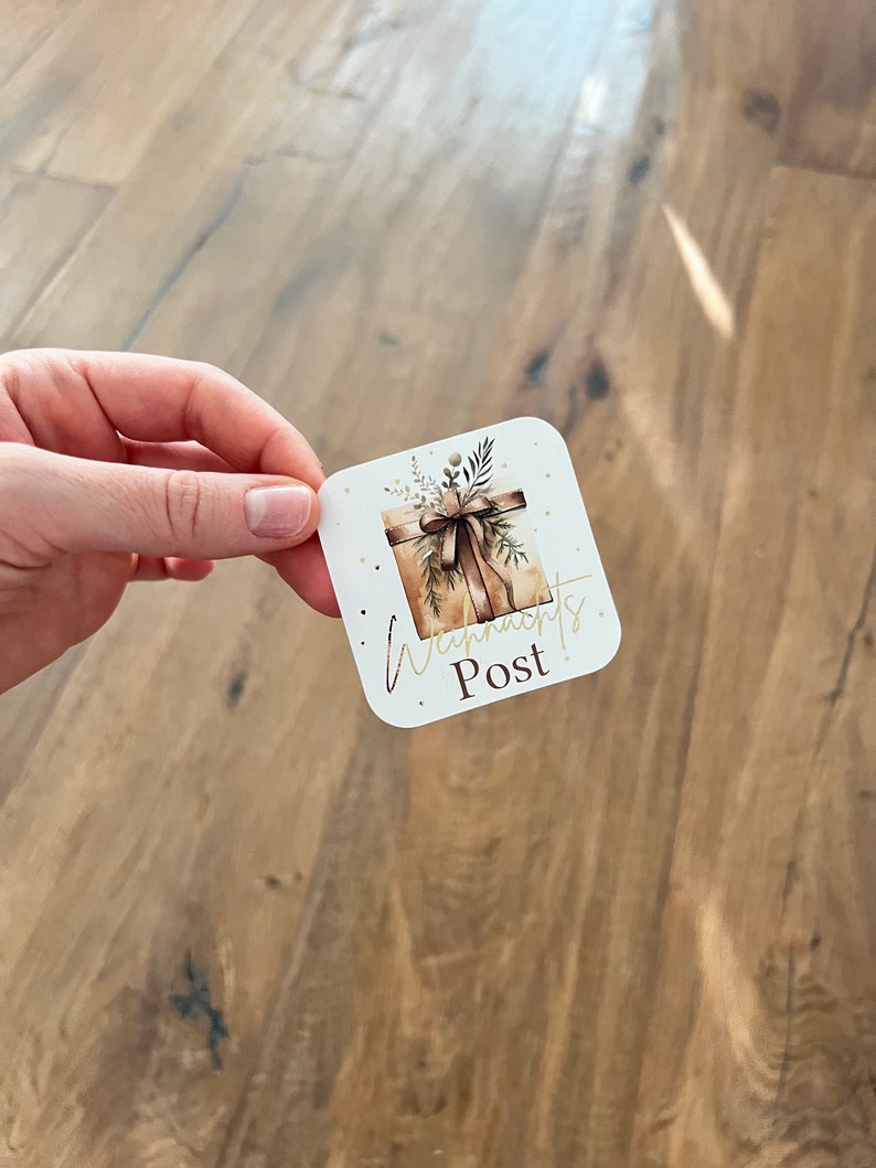 May include: A square sticker with rounded corners featuring a watercolor illustration of a wrapped gift box with a brown bow and greenery. The sticker has the words "Wynn's Post" in gold script.