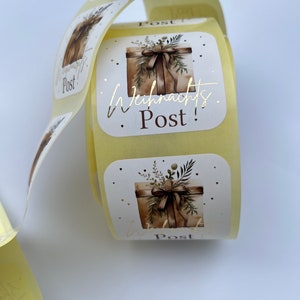 May include: A roll of square stickers with a watercolor illustration of a gift box tied with a ribbon and greenery. The text "Weihnachts Post!" is in gold foil. The stickers are on a roll with a light yellow backing.