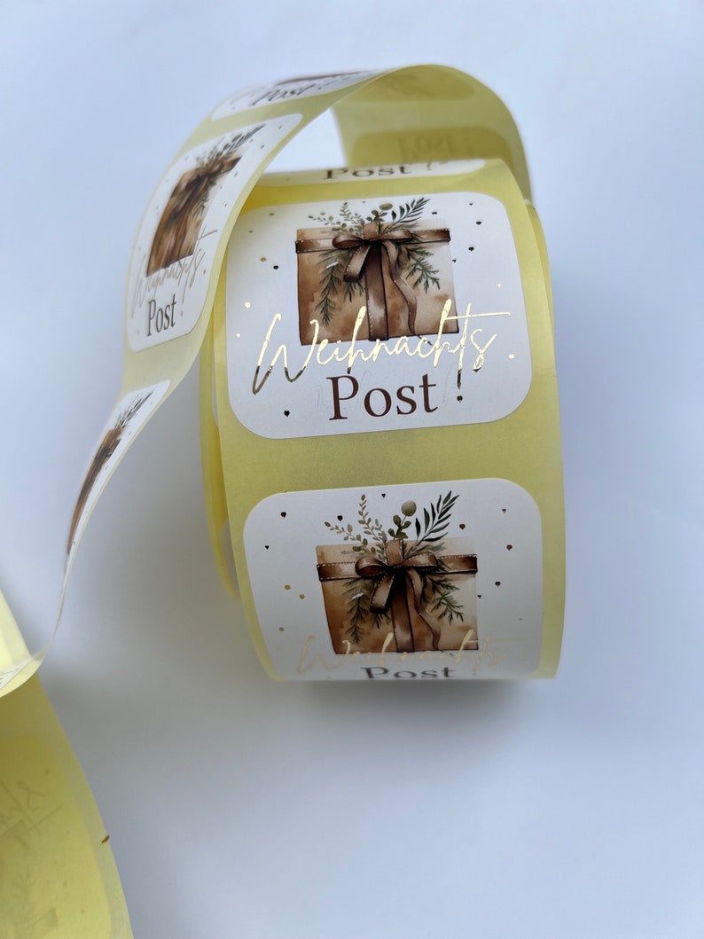 May include: A roll of square stickers with a watercolor illustration of a gift box tied with a ribbon and greenery. The text "Weihnachts Post!" is in gold foil. The stickers are on a roll with a light yellow backing.