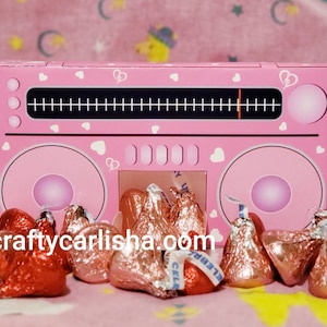 May include: Pink cardboard box shaped like a boombox with white hearts and a black dial. The box is open and filled with red and gold foil-wrapped chocolate kisses.