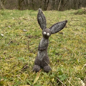 Puede incluir: Un conejo de fieltro gris con orejas largas y detalles blancos en la cara. El conejo está de pie sobre un lecho de hierba verde.