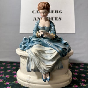 May include: A porcelain figurine of a woman in a blue dress sitting on a white pedestal. The woman is holding a small white object in her hands. The figurine is in a vintage style and has a delicate, feminine look.
