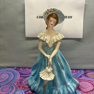 May include: A porcelain figurine of a woman in a long, light blue dress and a matching hat adorned with flowers. The figurine holds a small, white parasol. The text "CAR" and "QUES" is visible on a paper in the background.