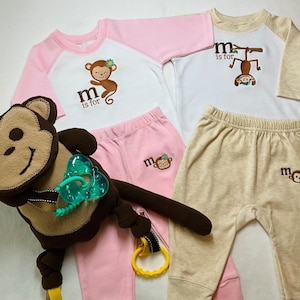 May include: A set of baby clothes with a monkey theme. The pink outfit includes a long-sleeved shirt with a white panel and the text "m is for" with a monkey graphic. The beige outfit includes a long-sleeved shirt with the text "m is for" and a monkey graphic. Both outfits include pants.