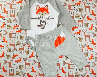 May include: A gray and white baby outfit with a fox design. The bodysuit has a white background with a red fox head and the text "Wild and Free". The pants are gray with a red fox tail on the back.