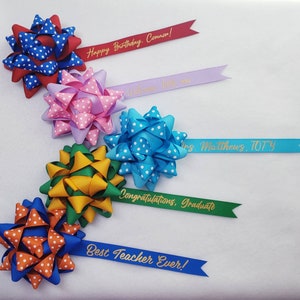 May include: Five colorful polka dot bows with gold lettering on attached ribbons. The ribbons read "Happy Birthday, Connor!", "Welcome little one", "Mrs. Matthews, TOTY", "Congratulations, Graduate", and "Best Teacher Ever!"