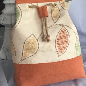 May include: A large, cream-colored drawstring bag with a brown cord and gold grommets. The bag is decorated with a pattern of colorful leaves. The bottom of the bag is a solid orange color.