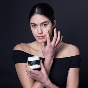 Model with glowing skin holds Naturally Wicked Citrus Charm Facial Serum, applying it to her cheek.