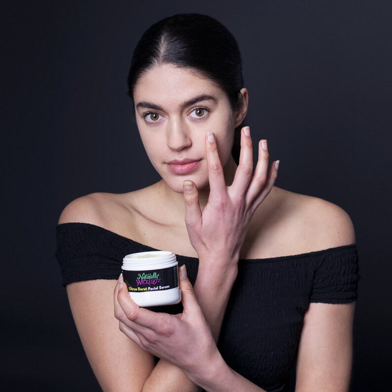 Model with glowing skin holds Naturally Wicked Citrus Charm Facial Serum, applying it to her cheek.