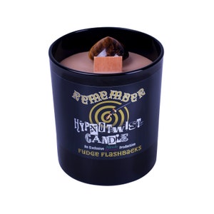 Pode incluir: Um frasco de vela preto com um rótulo dourado que diz "Remember HypnoTwist Candle An Exclusive Production Fudge Flashbacks". A vela tem uma parte superior de cera marrom e um cristal de olho de tigre no topo.