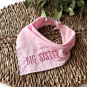 May include: Pink and white striped bandana with the embroidered text "BIG SISTER". The bandana is knotted at the top and is laying on a woven, brown surface. Eucalyptus leaves are in the background.