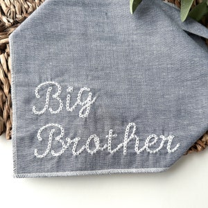 May include: A grey fabric bandana featuring the embroidered words "Big Brother" in white cursive. The bandana is folded and placed on a woven basket. The text is centered and stitched with a delicate, decorative pattern.