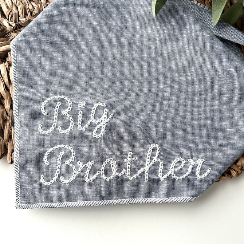 May include: A grey fabric bandana featuring the embroidered words "Big Brother" in white cursive. The bandana is folded and placed on a woven basket. The text is centered and stitched with a delicate, decorative pattern.