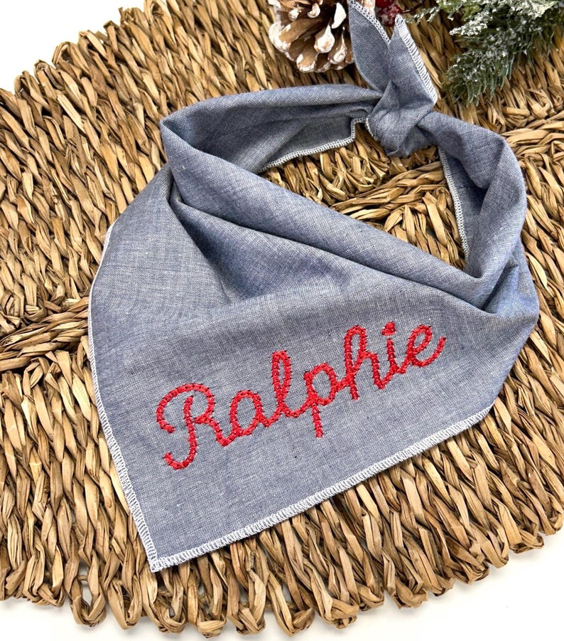 May include: A gray dog bandana with the name "Ralphie" embroidered in red thread. The bandana is made of a soft fabric and has a white stitched border. The background is a woven wicker material.