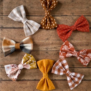 May include: Eight fabric bow ties arranged in a circle. Patterns include beige gingham, brown with white dots, rust with white crosses, plaid with brown and black, rust with white flowers, floral with mustard, solid mustard with crosses, and brown gingham.